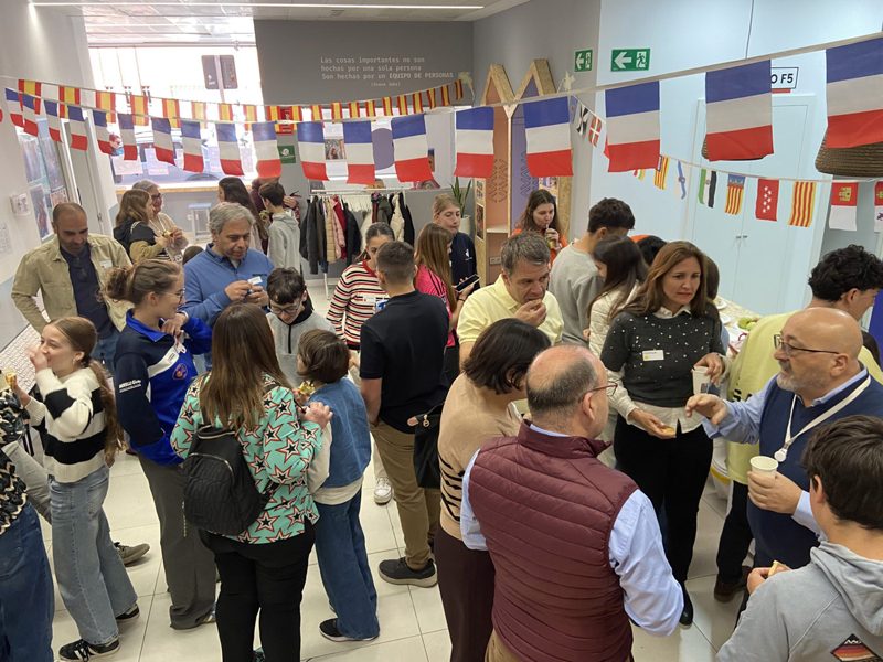 Familias y j&oacute;venes conversando y compartiendo comida en una sala decorada con banderas de Espa&ntilde;a, Francia y de las comunidades aut&oacute;nomas